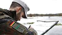 Ein Soldat trägt mit einem Stift etwas in eine Landkarte ein. Vor ihm erstreckt sich das Gelände.