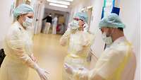 Zwei Frauen und ein Mann stehen mit Mundschutz, Kopfhaube und Kitteln bekleidet auf einem Krankenhausflur