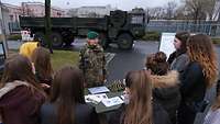 Soldatin vor einem militärischem Fahrzeug spricht zu einer Gruppe Schülerinnen.