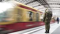 Eine Soldatin steht am Steig in einem Bahnhof. Eine S-Bahn fährt auf dem Gleis an ihr vorbei.