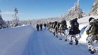 Soldaten laufen mit großen Rucksäcken und Schneeschuhen hintereinander durch eine Winterlandschaft.