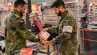 Zwei Soldaten stehen an einer Supermarktkasse, packen Sachen in einen Einkaufswagen 