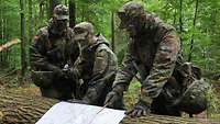Drei Soldaten mit Tarnschminke im Gesicht schauen auf eine Karte, die auf einem Baumstamm liegt.