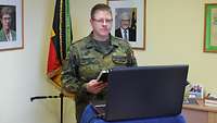 Ein Stabsoffizier steht in Uniform vor einem schwarzen Laptop in seinem Dienstzimmer.