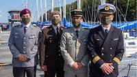 Vier Soldaten unterschiedlicher Nationen stehen am Bootshafen mit Mundschutzmasken vor Segelbooten und blicken in die Kamera.