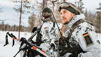 Zwei Soldaten stehen in Schneetarnanzügen, mit Schneestöcken und Waffen am Rand eines Waldes.