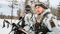 Zwei Soldaten stehen in Schneetarnanzügen, mit Schneestöcken und Waffen am Rand eines Waldes.