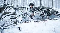 Ein Soldat in Schneetarn baut aus Holzstämmen und Planen einen Unterstand im Schnee.