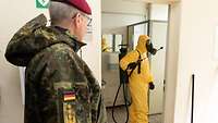 Soldat sieht durch eine Türöffnung, wie eine Person mit Schutzanzug und Maske Desinfektionsmittel versprüht.