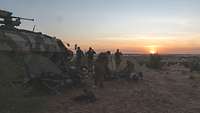 Die Soldaten bauen ihre Feldbetten ab, daneben ein Transportpanzer. Am Horizont geht die Sonne auf.