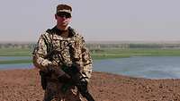 Ein Soldat in Tropentarn mit Waffe steht auf sandigem Boden, hinter Grünflächen und der Fluß Niger.