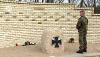 Ein Soldat steht vor einer Steinwand mit kleinen Tafeln, neben ihm ein Gedenkstein mit dem Eisernen Kreuz