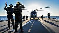 Auf dem Flugdeck eines Kriegsschiffs steht ein Hubschrauber, mehrere Marinesoldaten stehen daneben.