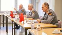 Drei Männer mit grauen Haaren in grauer Uniform sitzen mit Abstand nebeneinander an Konferenztischen.