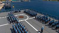 Aus der Vogelperspektive: Marinesoldaten in blauer Arbeitsuniform stehen in geraden Reihen auf dem Flugdeck eines Kriegsschiffs.