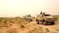 In einer Wüste fahren mehrere Militärfahrzeuge auf einem sandigen Weg hintereinander.