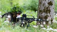 Ein Soldat mit Mund-Nasenschutz und getarntem Helm liegt mit einem Gewehr im Anschlag in einer Wiese