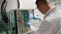 Ein Mann in weißen Kittel mit Handschuhen und Brille arbeitet an einer Laborapperatur