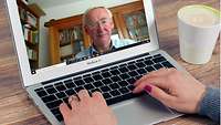 Ein Mann mit grauen Haaren und Brille ist auf dem Bildschirm eines Laptops zu sehen. Am Laptop Frauenhände, daneben eine Tasse K