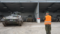 Ein Soldat mit orangener Warnweste von hinten, vor ihm ein Panzer