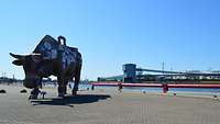 Eine graue Hafenpier auf der ein Denkmal in Form einer Kuh steht, im Hintergrund Industrieanlagen.