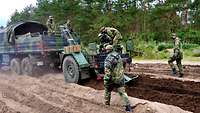 Der Anhänger an einem Lkw pflügt den Boden. Soldaten bedienen das Gerät und laufen hinter ihm her.