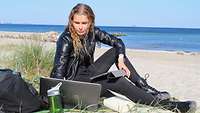 Eine junge Frau mit blonden Locken sitzt mit ihrem Laptop am Strand.