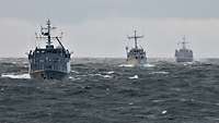 Drei kleine graue Kriegsschiffe von vorne in rauher See.