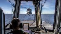 Blick durch drei Fenster auf das Flugdeck eines Schiffes in See, über dem ein Hubschrauber schwebt.