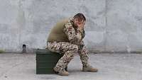 Ein Soldat sitzt auf einer Kiste und hält sich verzweifelt die Hände vor das Gesicht
