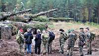 Neun Soldaten und zwei Journalisten stehen neben Panzern auf dem Übungsplatz.