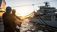 Zwei Marinesoldaten in dunkelblauer Arbeitsuniform und Schutzhelmen auf Deck eines Schiffs, im Hintergrund rein zweites Schiff.