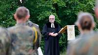 Pfarrer Johannes Müller steht im Talar an einem Stehpult, das als Kanzel dient. Im Vordergrund stehen Soldaten.