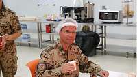 In der Küche sitzt der Militärpfarrer mit Haarnetz vor einem Dessert, er hält einen Kaffeebecher in der Hand