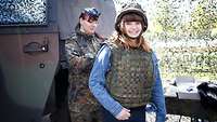 Ein Mädchen mit Helm und Schützweste lächelt in die Kamera, eine Soldatin neben einem Militärfahrzeug im Hintergrund