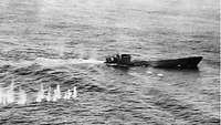 Schwarzweiß-Aufnahme eines U-Boots an der Wasseroberfläche; im Wasser weiße Spitzer von einschlagenden Geschossen.