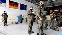 Soldaten mit Helmen und Fallschirmen stehen in einem Hangar. An der Wand hängen drei Flaggen.