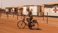 Ein Soldat fährt auf einer Wüstenstraße Fahrrad, im Hintergrund Container und Zelte