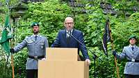 Ein Mann in einem Anzug steht hinter einem Rednerpult, dahinter stehen Soldaten mit Fahnen.