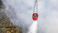 Ein an einem Hubschrauber befestigter Wasserbehälter wird in der Luft geöffnet und löscht so einen Brand
