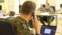 Ein Soldat sitzt in der Operationszentrale an einem Schreibtisch und telefoniert. Im Hintergrund weitere Soldaten.