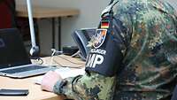 Ein Feldjäger sitzt an einem Schreibtisch der mit Laptop und Telefon versehen ist.