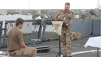 Ein Militärpfarrer predigt auf dem Flugdeck der deutschen UNIFIL-Korvette, links auf einer Bank sitzt ein Soldat