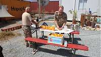 Ein Militärpfarrer presst Orangensaft, auf dem Tisch stehen einige Kannen, auf einer Sitzbank eine Kiste Orangen