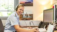 Eine Soldatin in hellblauem Uniformhemd mit Headset ist von der Seite vor ihrem PC zu sehen. Sie lächelt in die Kamera.