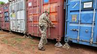 Ein Soldat steht vor mehreren Containern in verschiedenen Farben, seine Hände umfassen die Verriegelung eines Containers 