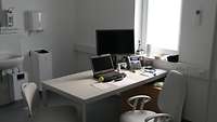 Ein Arbeitszimmer für den Arzt mit Laptop, großem Bildschirm und Telefon, im Hintergrund ein Waschbecken