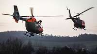 Zwei grün-orangefarbene Hubschrauber überfliegen in geringer Höhe ein Dorf in der Dämmerung.