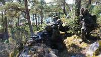 In einem Waldstück warten Soldaten in Flecktarn und Gewehren in der Hand. 