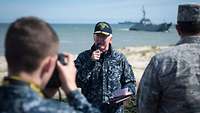 Ein Soldat in blau-grauem Flecktarn steht am Strand und spricht in ein Mikrofon.